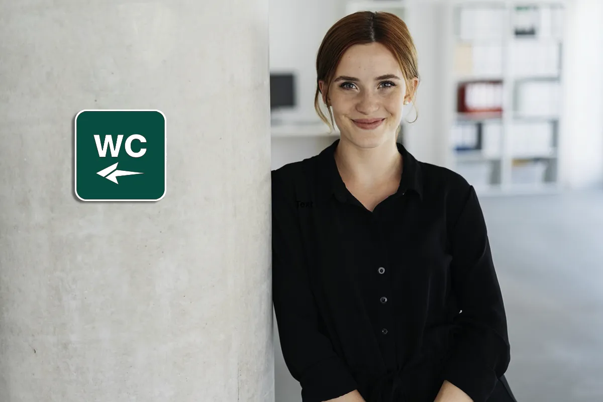Eine weibliche Person steht gegen eine Wand gelehnt neben einem grünen WC-Schild mit einem Pfeil nach links